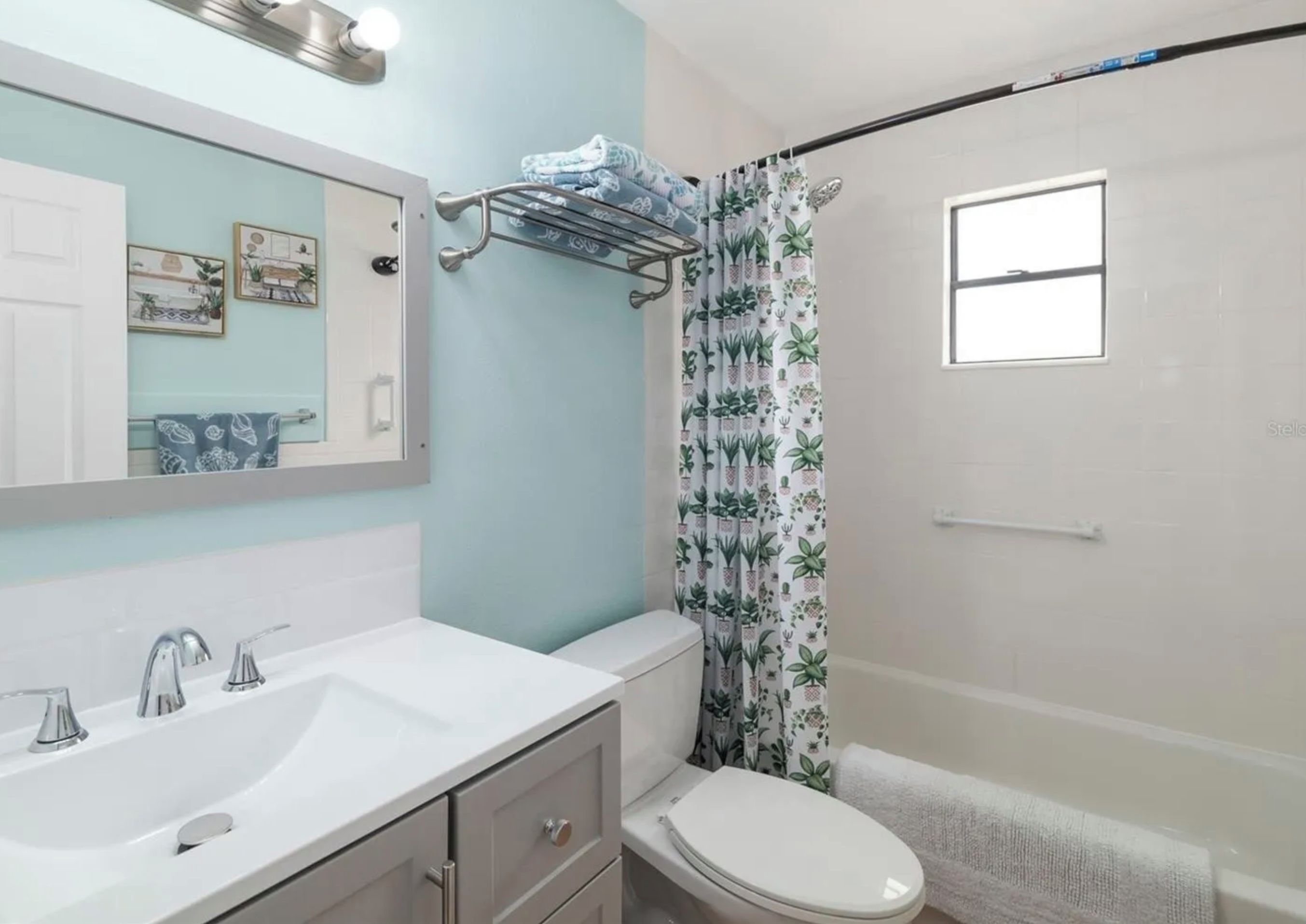 Une salle de bain avec un lavabo blanc, des toilettes, un rideau de douche orné d’un motif végétal et un miroir agrémenté d’une décoration murale.