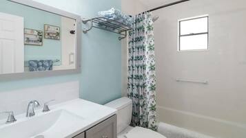 Une salle de bain avec un lavabo blanc, des toilettes, un rideau de douche orné d’un motif végétal et un miroir agrémenté d’une décoration murale.