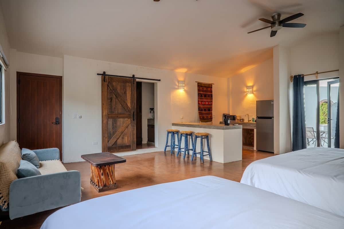 A modern hotel room with a large bed, a small kitchen area, and a sliding door leading to a balcony.