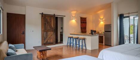 A modern hotel room with a large bed, a small kitchen area, and a sliding door leading to a balcony.