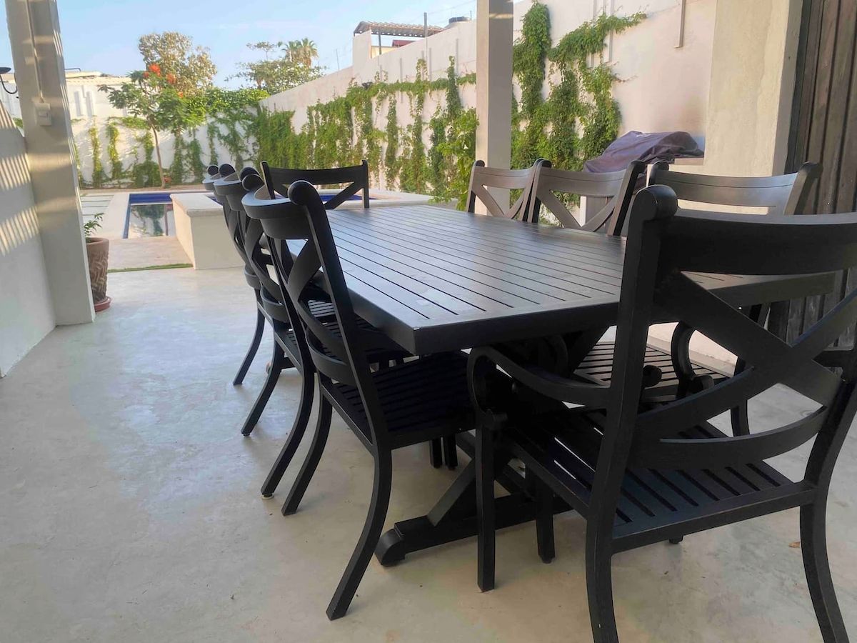 Une terrasse extérieure avec une table rectangulaire et six chaises, entourée d’un sol en béton et d’un petit muret agrémenté de verdure.