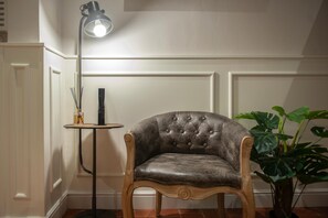 Een gestoffeerde fauteuil met houten armleuningen en een klein bijzettafeltje met een lamp, geplaatst in een kamer met witgekleurde wandpanelen en een potplant.