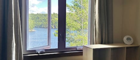 Une pièce avec une fenêtre donnant sur un lac et une forêt, une étagère en bois et un objet blanc posé sur l’étagère.