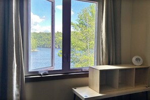 Une pièce avec une fenêtre donnant sur un lac et une forêt, une étagère en bois et un objet blanc posé sur l’étagère.