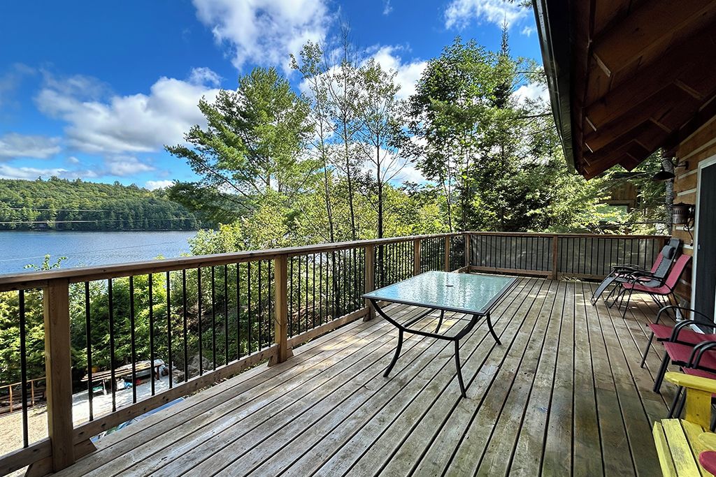 Una terraza de madera con una mesa y sillas de vidrio, con vistas a un lago y un bosque.