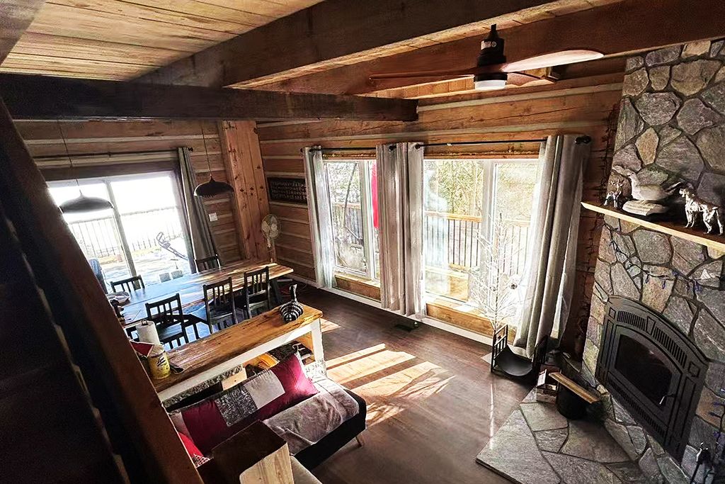 A rustic cabin interior with a stone fireplace, wooden beams, a dining area, and large windows.
