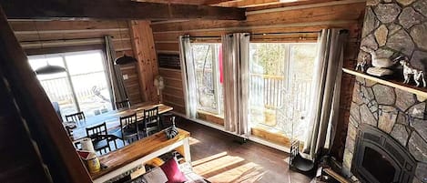 A rustic cabin interior with a stone fireplace, wooden beams, a dining area, and large windows.