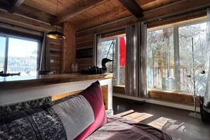 Interior de una cabaña con techo de madera, una zona de bar, un sofá con cojines y ventanas grandes con cortinas.