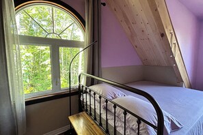Un lit simple avec un cadre en métal et du linge de lit blanc, un banc en bois et une fenêtre donnant sur des arbres.