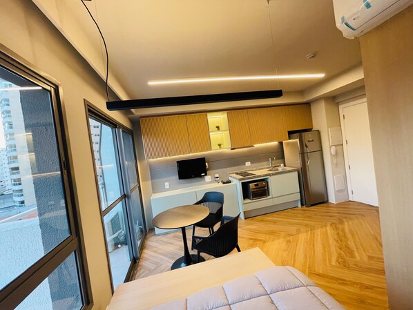 Ein modernes Hotelzimmer mit großem Fenster, einem Bett, einem kleinen Esstisch und einer Küchenzeile.