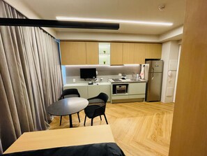 Quarto de hotel compacto com cozinha compacta, área de refeições com mesa e cadeiras, e uma janela grande com cortinas.