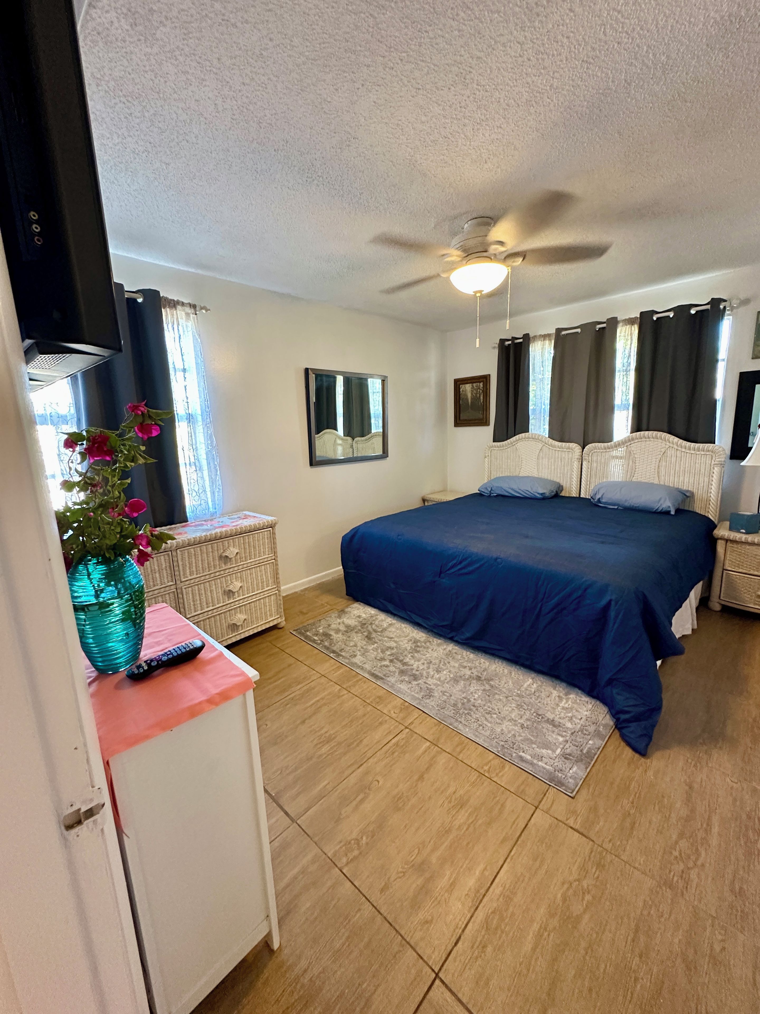 A bedroom with a bed, a ceiling fan, a dresser, a vase with flowers, and a window with curtains.