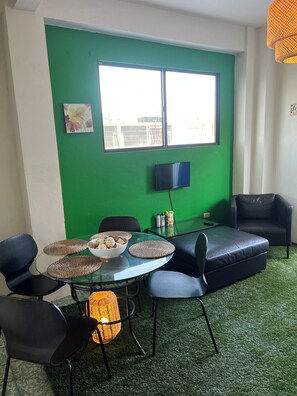 Et værelse med en grøn væg, et tv ophængt på væggen, et glasbord med vævede underlag, en skål med snacks og en sort lædersofa.