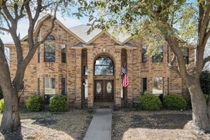 A two-story brick building with a central entrance, an American flag, and a symmetrical facade. - Light-filled 6-bedroom furnished home, 3,300 sqft, monthly stays (Plano)