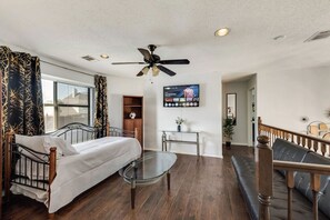 A bedroom with a bed, a glass coffee table, a ceiling fan, a TV, and a view of the outside. - Light-filled 6-bedroom furnished home, 3,300 sqft, monthly stays (Plano)