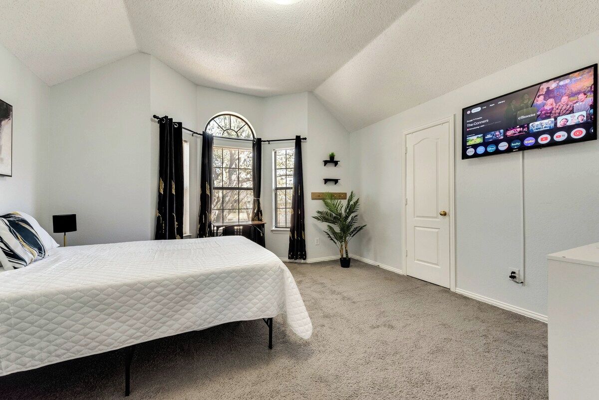 A bedroom with a bed, a TV mounted on the wall, a window with curtains, and a door.
