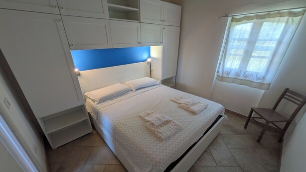 A bedroom with a bed, built-in cabinets, a chair, and a window with curtains. - Apartment just a few meters from the beach (Quartu Sant'Elena)