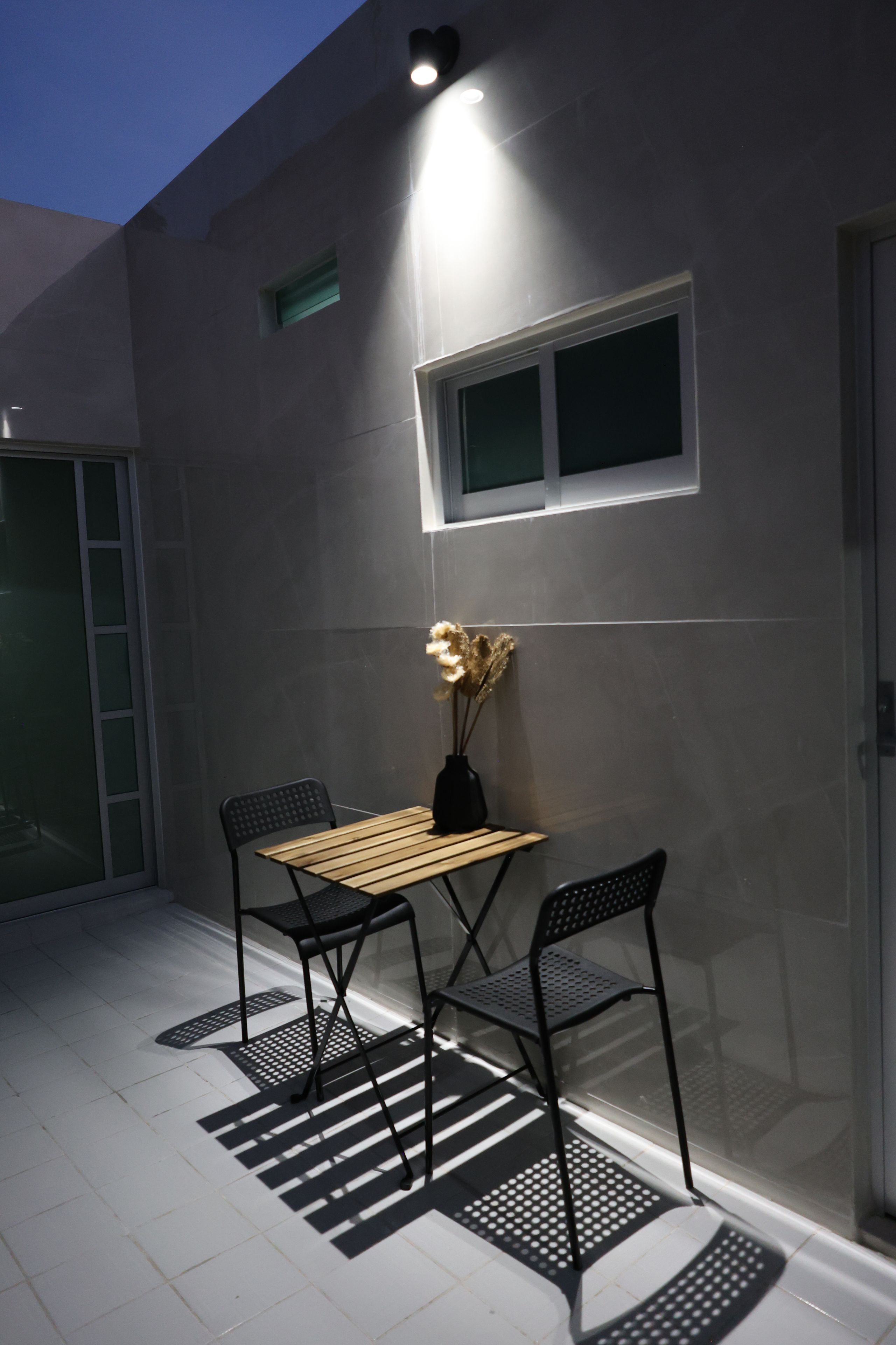 A patio with a table and chairs, a vase with dried flowers, and a window with a light fixture.