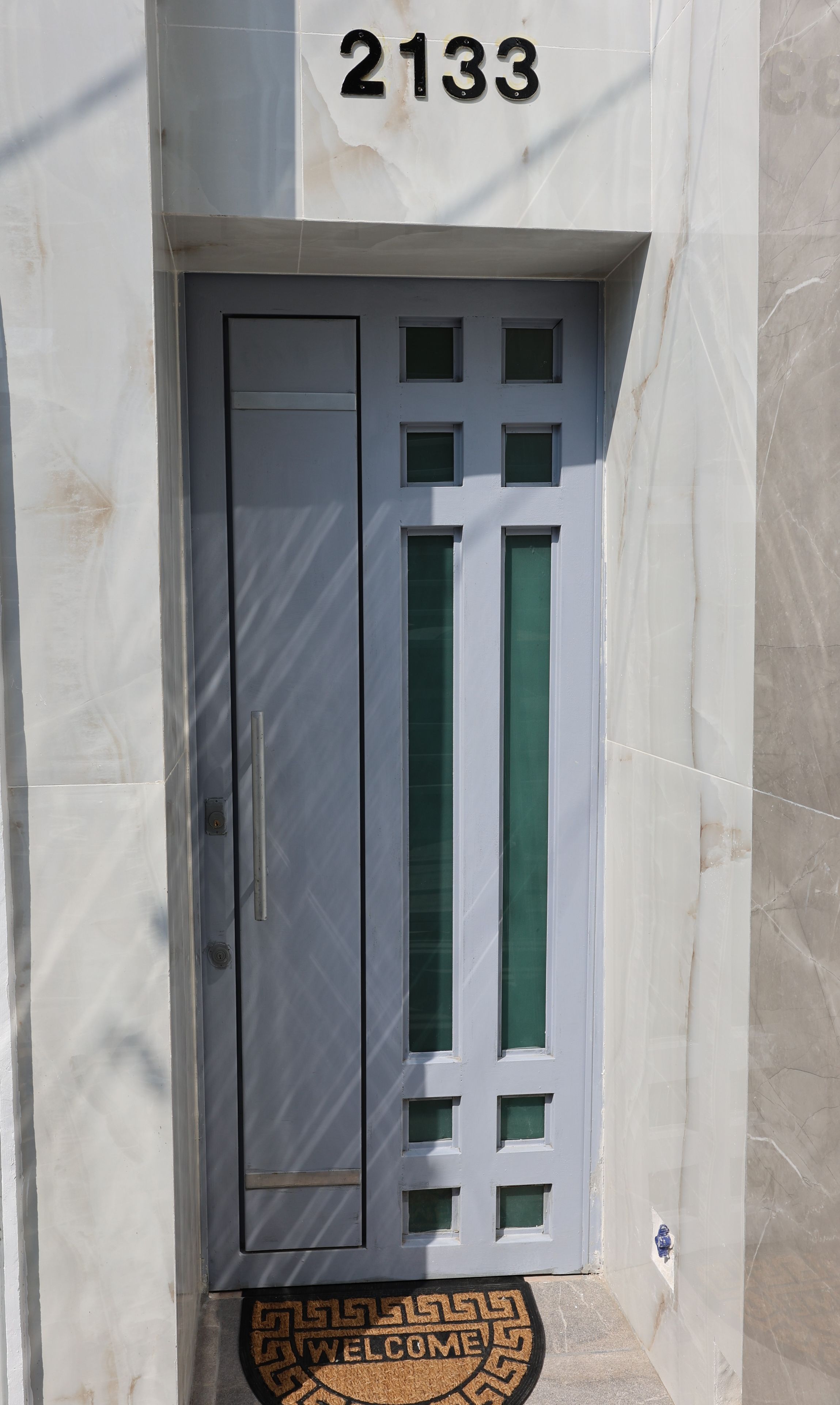 A white door with a number '2133' above it, a doormat with 'WELCOME' and a keyhole, and a small window with a green-tinted glass.