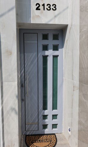 A white door with a number '2133' above it, a doormat with 'WELCOME' and a keyhole, and a small window with a green-tinted glass.