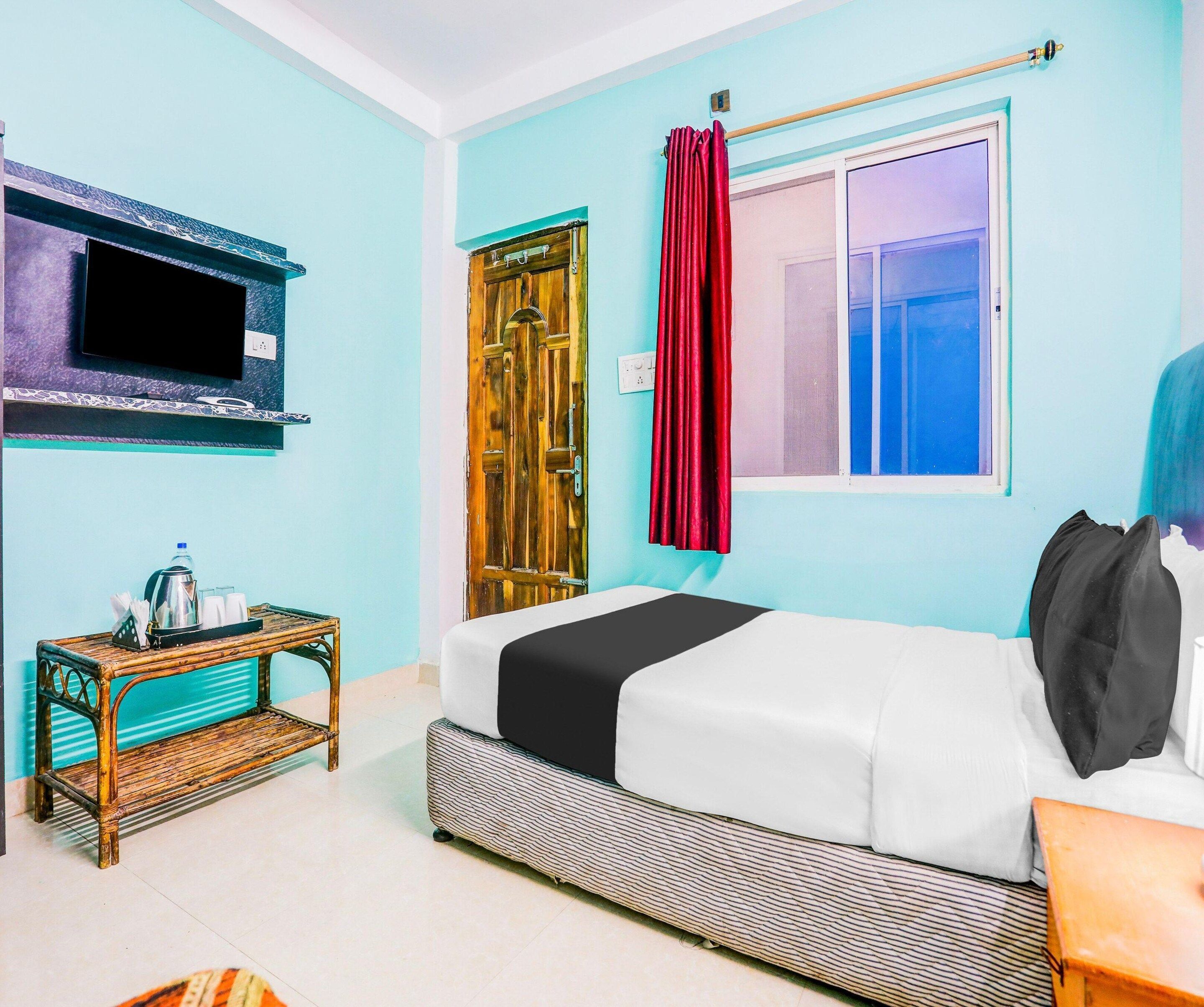 A room with a bed, a television mounted on the wall, a window with curtains, and a small table with a water bottle.