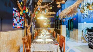 A restaurant interior with a long dining table, wooden chairs, hanging bamboo baskets, and colorful hanging bells.