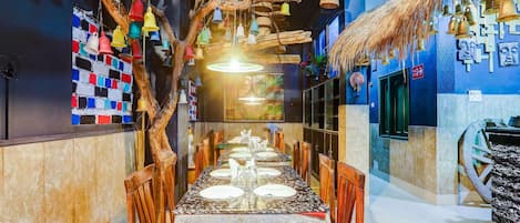 A restaurant interior with a long dining table, wooden chairs, hanging bamboo baskets, and colorful hanging bells.