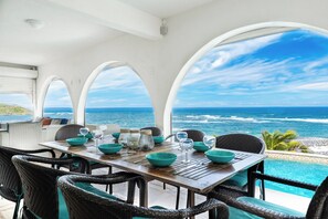 An outdoor dining area with a view of the ocean, featuring a wooden table and wicker chairs. - Villa Oyster Pearl: Concierge Hot Tub Beach Access (Oyster Pond)