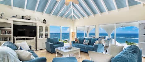 A spacious living room with a view of the ocean, featuring a ceiling fan, a television, and a bookshelf.