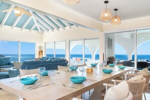 A spacious dining area with a long table set for six, large windows offering ocean views, and a ceiling fan with a light fixture. - Villa Oyster Pearl: Concierge Hot Tub Beach Access (Oyster Pond)