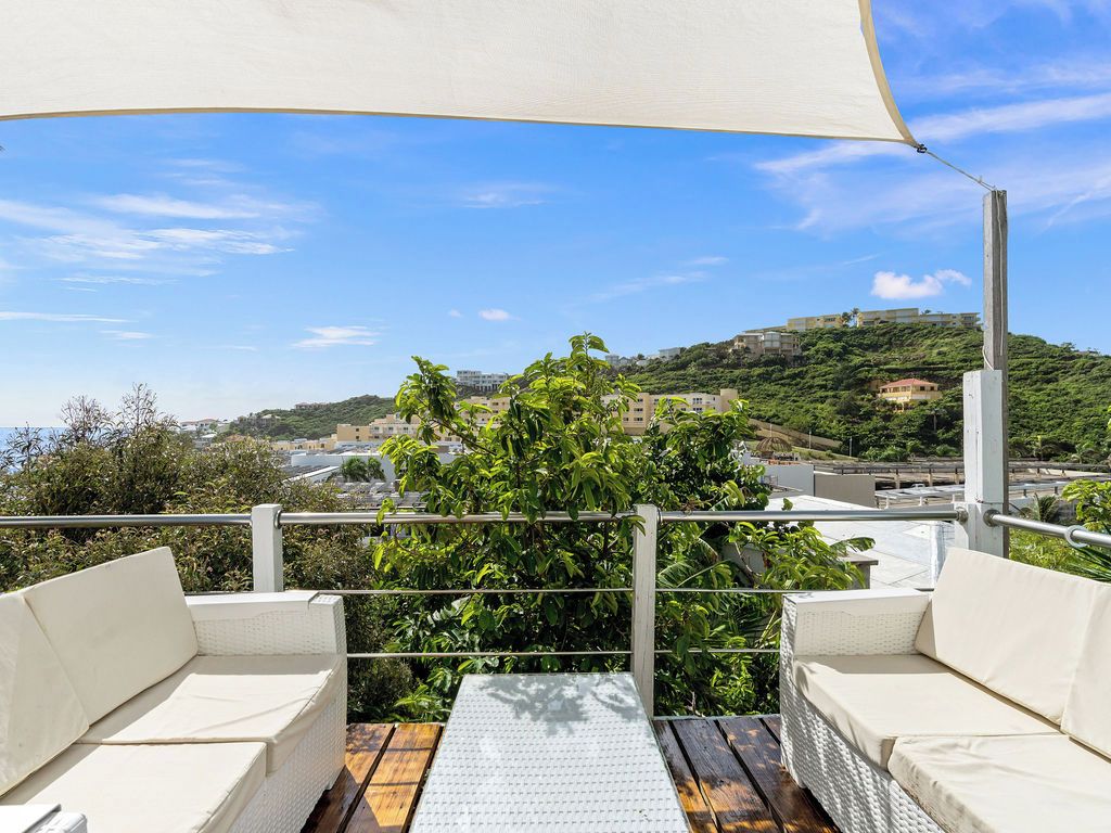 A terrace with white cushioned seating and a wooden deck, overlooking a hillside with buildings and greenery.