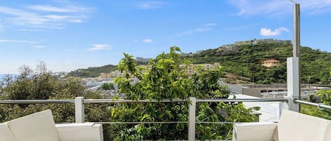A terrace with white cushioned seating and a wooden deck, overlooking a hillside with buildings and greenery.