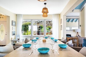 A dining area with a long table set for six, surrounded by wicker furniture, and a view of a garden through large windows. - Villa Oyster Pearl: Concierge Hot Tub Beach Access (Oyster Pond)