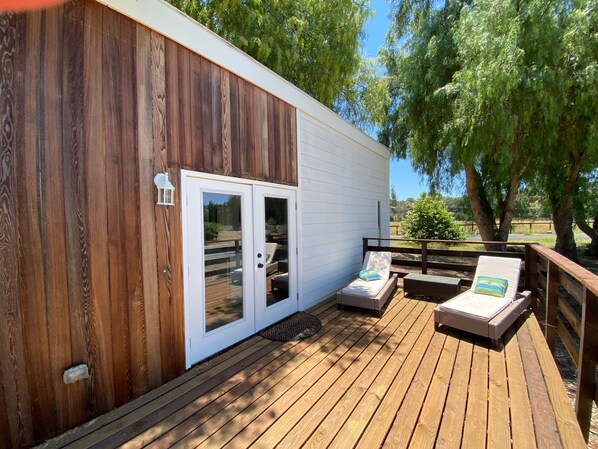 A wooden deck with two lounge chairs and a small table, a white door with glass panels, and a lantern on the wall.