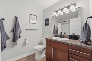 Une salle de bain moderne avec des toilettes, un lavabo et un meuble-lavabo. Des serviettes sont suspendues à des crochets et un tableau encadré est accroché au mur.