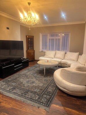 Ein Wohnzimmer mit einem großen Flachbildfernseher, einem runden Couchtisch, einem beigen Sofa, einem gemusterten Teppich und einer Chandelieber.
