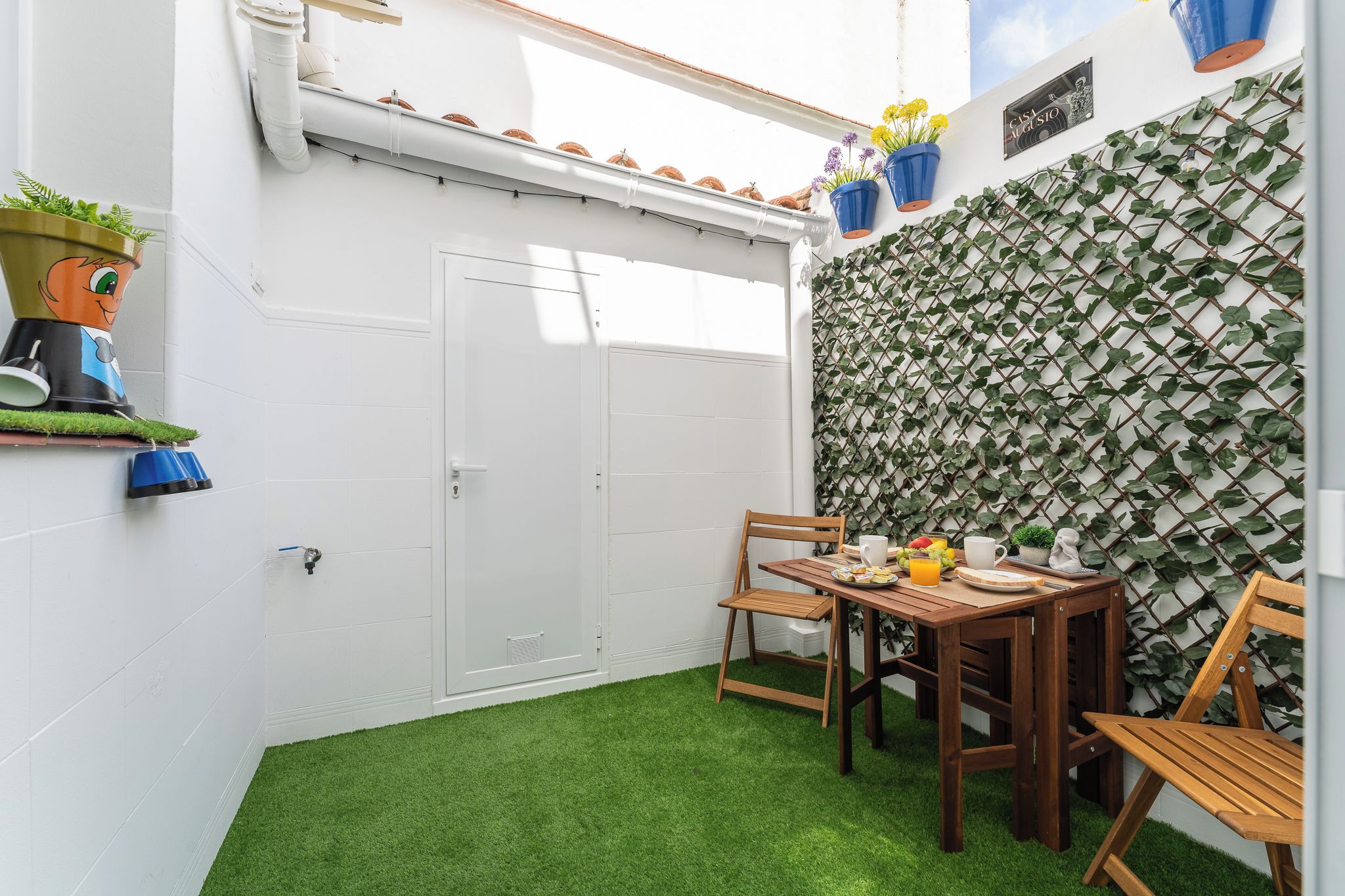 Un pequeño patio con una mesa de madera dispuesta para el desayuno, una puerta blanca y una pared cubierta de plantas trepadoras.
