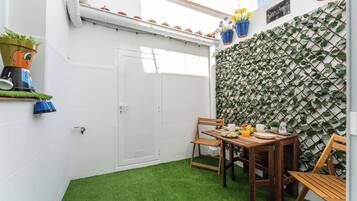 Un pequeño patio con una mesa de madera dispuesta para el desayuno, una puerta blanca y una pared cubierta de plantas trepadoras.