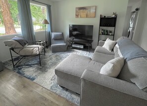 A modern living room with a grey sofa, a black TV stand, a small table, and a bookshelf. - The Raleigh Residence — 5 min to Downtown (Raleigh)