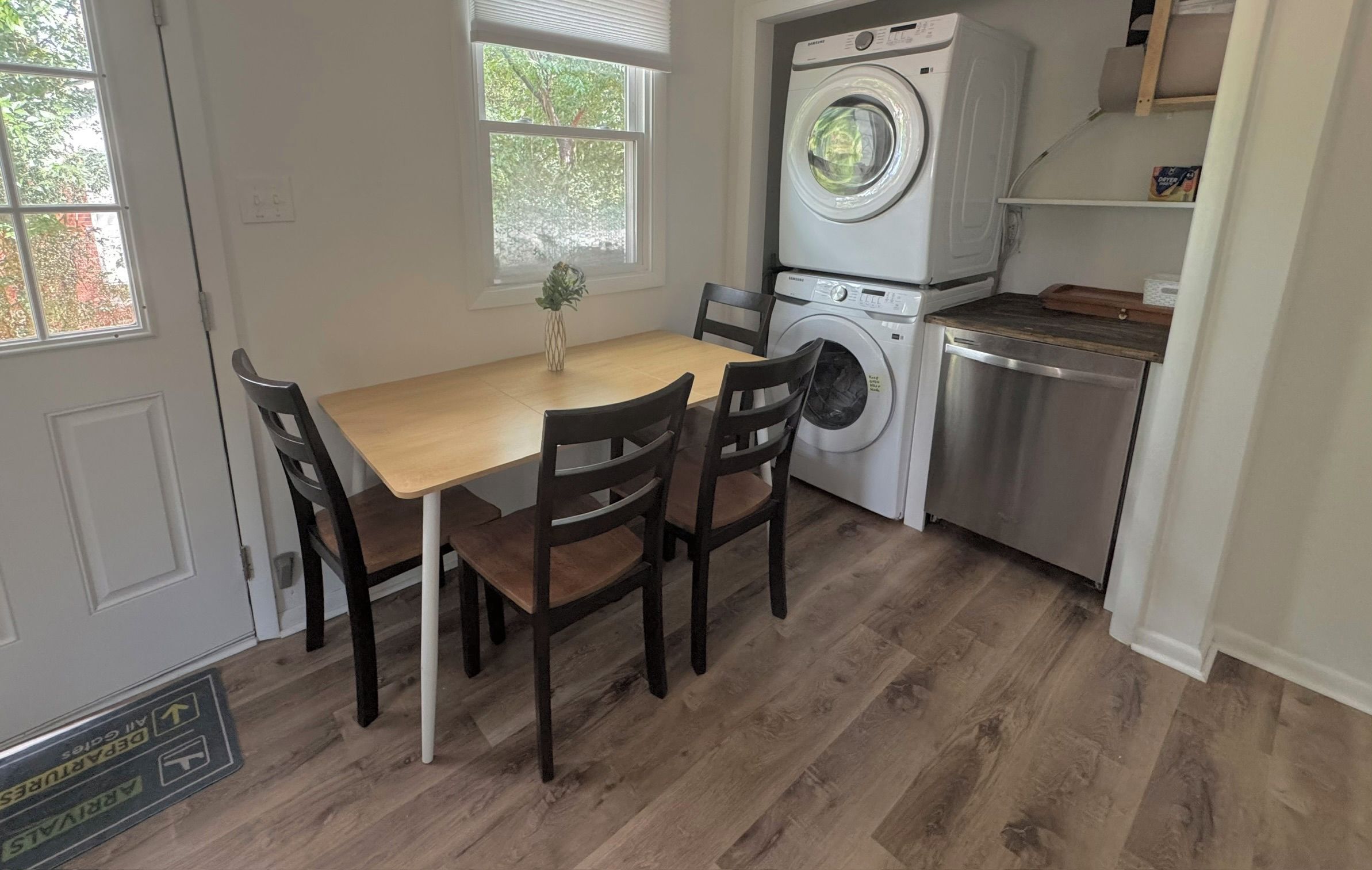 Una cocina funcional con lavadora, lavavajillas, una mesa de comedor con sillas y una ventana con vistas a árboles.