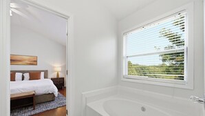 Un baño moderno con una bañera independiente, una ventana con persianas, y se ve un dormitorio con cama y mesita de noche.