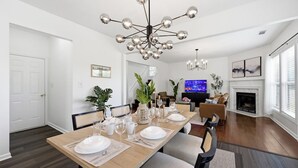 Un comedor con mesa de madera dispuesta para cuatro, una lámpara moderna y una sala de estar con chimenea.