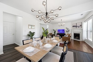 Uma sala de jantar com mesa de madeira posta para quatro pessoas, lustre moderno e uma sala de estar com lareira.