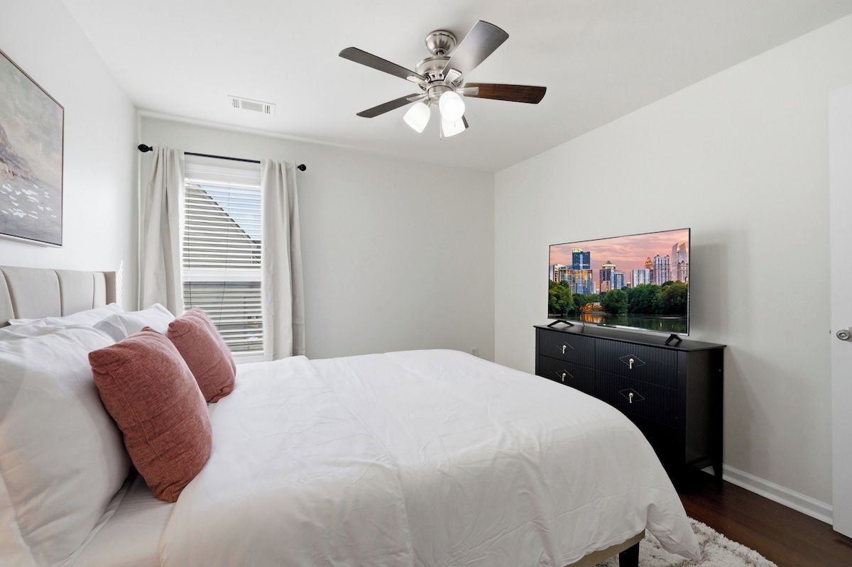 A bedroom with a bed, a ceiling fan, a TV on a dresser, and a window with curtains.
