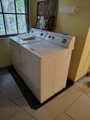 A laundry area with a front-loading washer and dryer, a window with a view of trees, and a painting on the wall. - La Finca 1928 w stream in Valley of Moon/Historical 1 Bd 2Bath two-level cabin (Crestline)