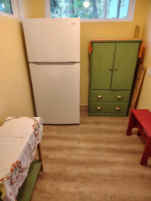 Eine kleine Küche mit einem weißen Kühlschrank, einem grünen Schrank mit Schubladen, einer Holzbank und einem kleinen roten Hocker.