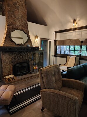 A cozy living room with a stone fireplace, a comfortable armchair, and a view of the outdoors through a window. - La Finca 1928 w stream in Valley of Moon/Historical 1 Bd 2Bath two-level cabin (Crestline)
