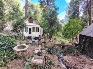 A rustic cabin surrounded by dense forest, with a small outdoor seating area and a visible window. - La Finca 1928 w stream in Valley of Moon/Historical 1 Bd 2Bath two-level cabin (Crestline)