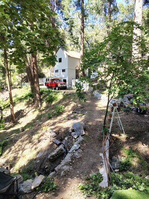 A white house with a red truck parked outside, surrounded by trees and a dirt path. - La Finca 1928 w stream in Valley of Moon/Historical 1 Bd 2Bath two-level cabin (Crestline)