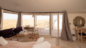 A room with a large bed, a sofa, a dining table, and chairs. There are large windows with a view of a desert landscape.